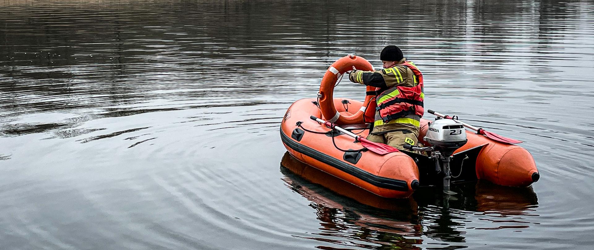Zdjęcie wykonano w porze dziennej przy pochmurnym niebie. Na wodzie znajduje się pomarańczowy ponton ratunkowy wyposażony w silnik oraz wiosła. Na pontonie znajduje się strażak ubrany w ubranie specjalne i kamizelkę ratunkową, który przygotowuje się do rzutu koła ratunkowego. W tle znajduje się woda oraz drzewa. 