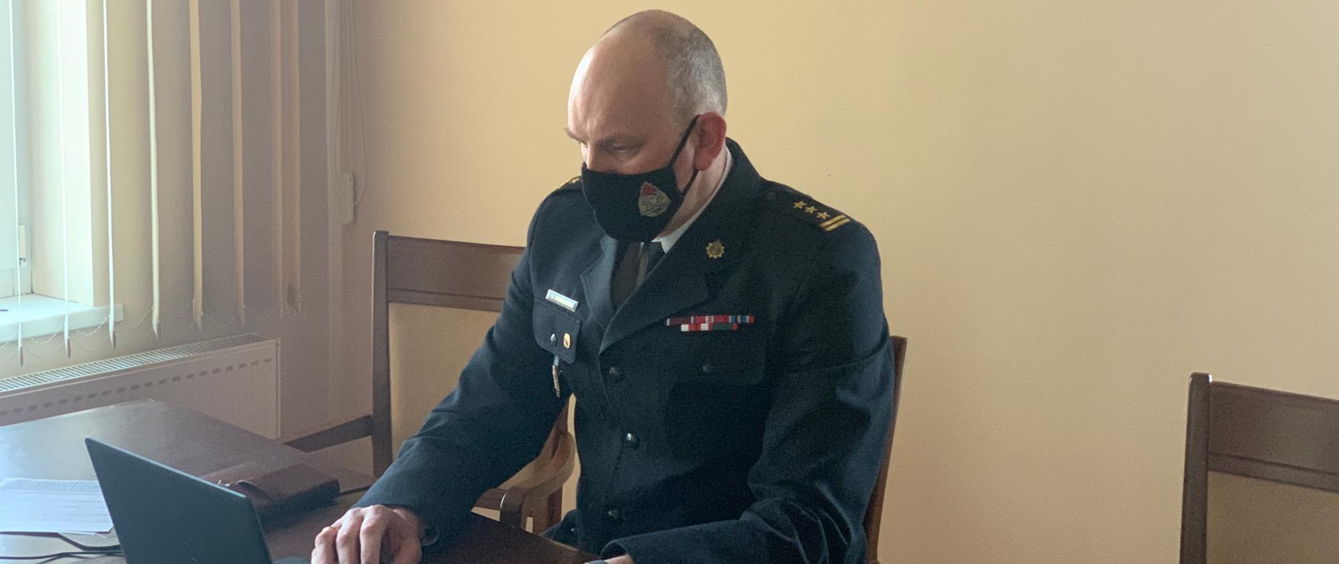 Na zdjęciu widać strażaka w umundurowaniu wyjściowym. Siedzi on przy biurku przed laptopem, nad nim na ścianie logo PSP i napis Komenda Wojewódzka Państwowej Straży Pożarnej w Toruniu.