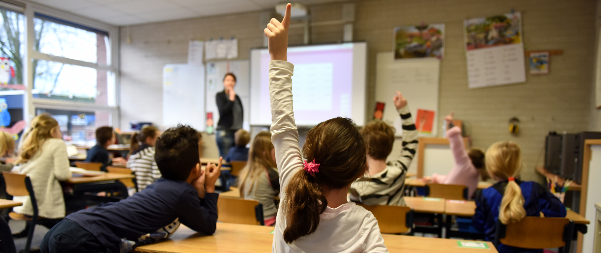 Na pierwszym planie dziewczynka w białej bluzce z długim rękawkiem, z ciemnymi włosami związanymi w kitkę zgłasza się do odpowiedzi, obok chłopiec oparty łokciami na ławce. w tle dzieci siedzące w ławkach, przy tablicy nauczycielka. 