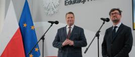 Minister Czarnek i mężczyzna w czarnym garniturze stoją za mikrofonami, za nimi flagi Polski i UE i napis na ścianie Ministerstwo Edukacji i Nauki.