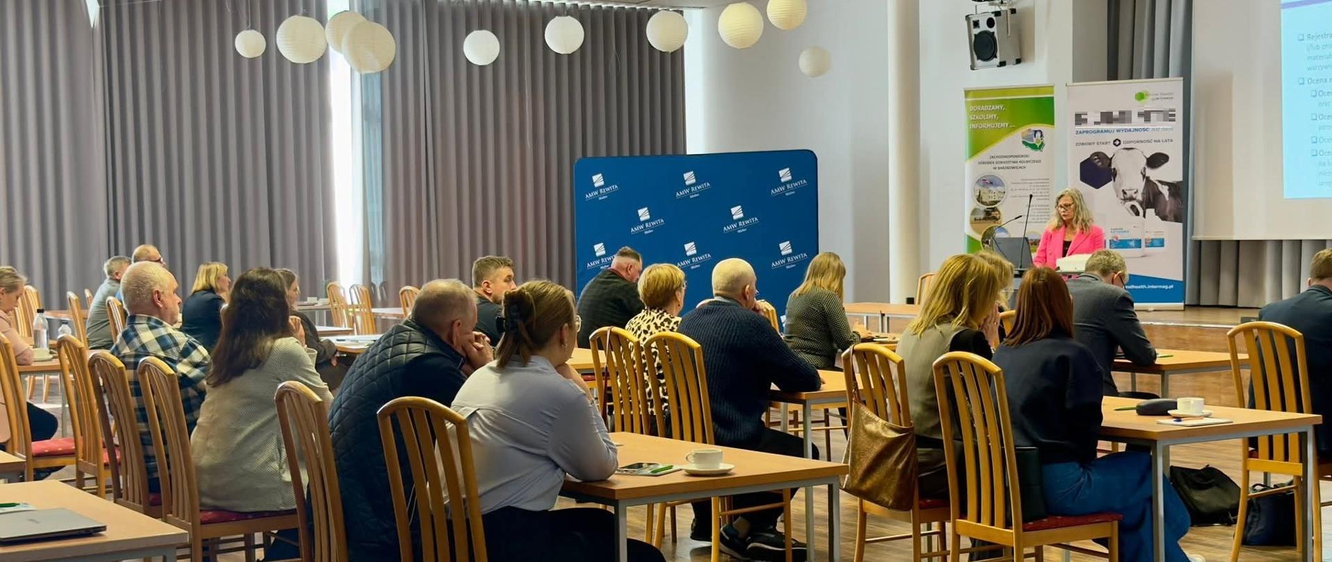 Zdjęcie przedstawia salę wypełnioną ludźmi siedzącymi w szkolnych ławkach. Po prawej stronie można zobaczyć część prezentacji wyświetlonej na ekranie projekcyjnym.