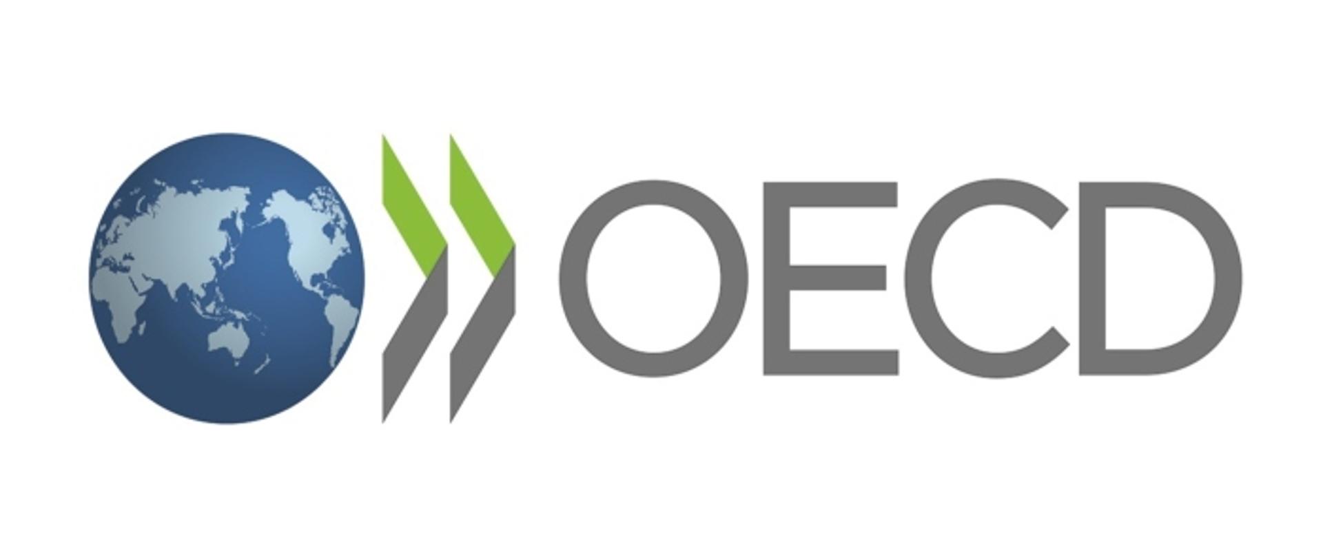 Logo OECD. Od lewej rysunek kuli ziemskiej w barwach błękitnych, kontynenty zaznaczone jaśniejszym odcieniem. Dalej w prawo dwa zielono-szare elementy graficzne, przypominające otwarte okładki książek, skierowane grzbietami w prawo. Dalej w prawo napis szarymi literami: OECD.