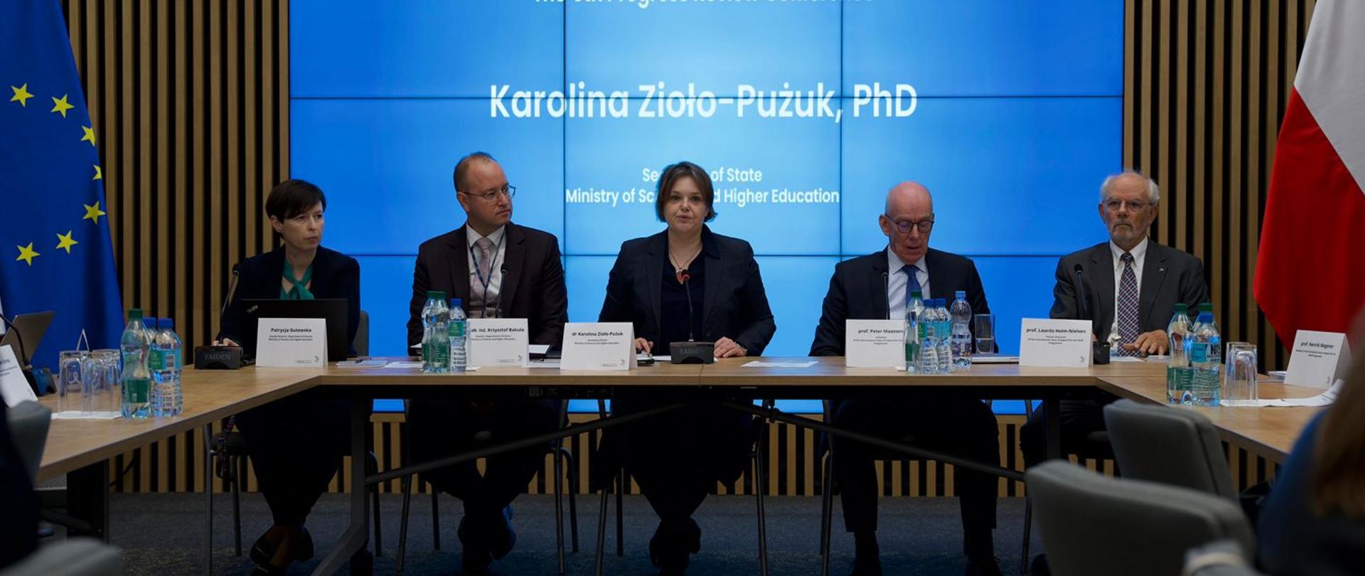 Zdjęcie od tyłu sali, za stołem siedzi trzech mężczyzn i dwie kobiety, pośrodku wiceminister Zioło-Pużuk, za nimi na wbudowanym w ścianę wielkim niebieskim ekranie napis Karolina Zioło-Pużuk PhD.