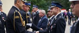 Zdjęcie zrobione na zewnątrz, podczas uroczystego apelu. Oficer Państwowej Straży Pożarnej w stopnia starszego brygadiera wręczając oznaczenie podaje dłoń strażakowi ochotnikowi.
