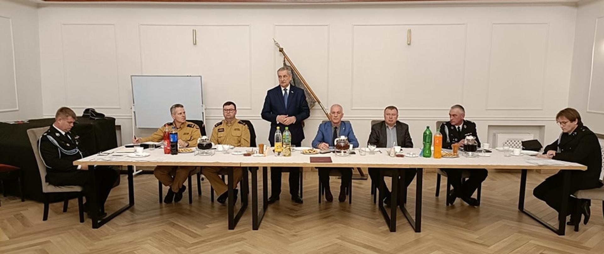 Strażacy w mundurach i osoby cywilne siedzą na sali przy stole.