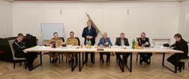 Strażacy w mundurach i osoby cywilne siedzą na sali przy stole.