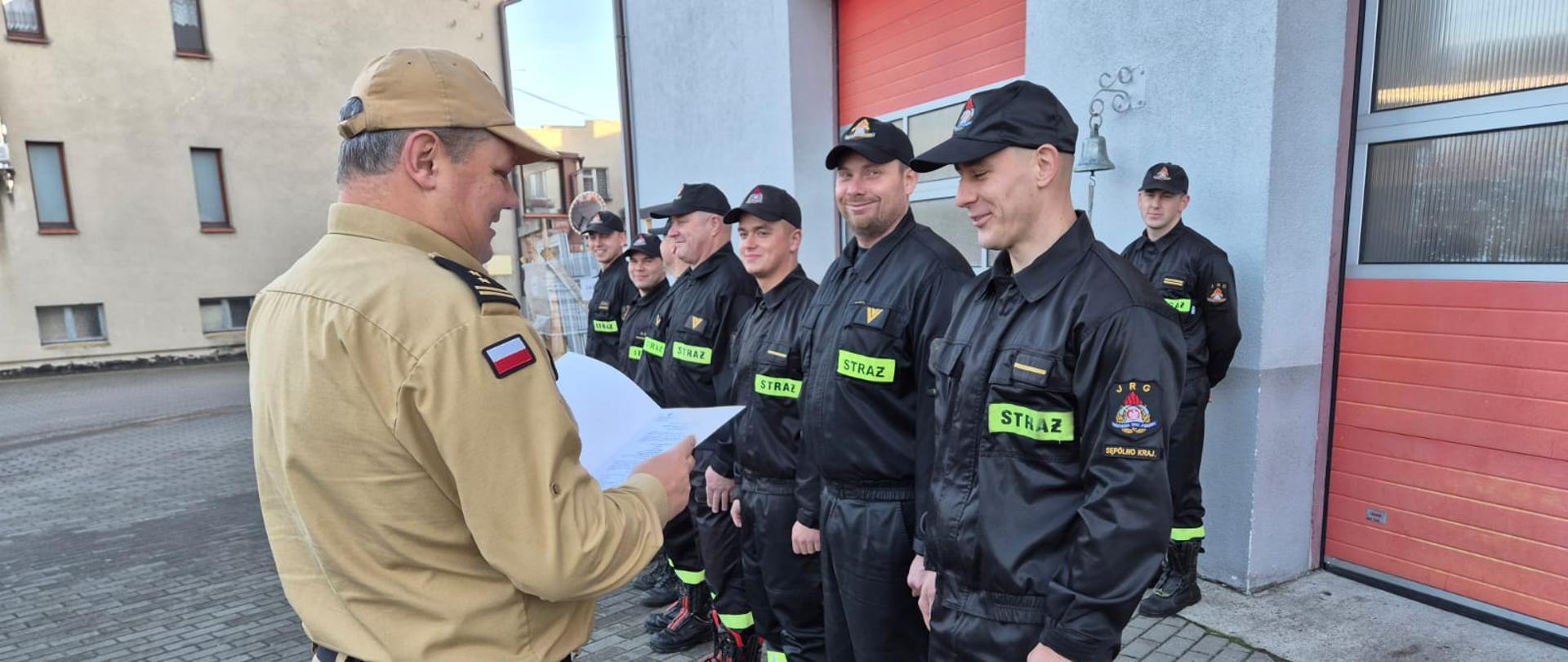 Plac Komendy Powiatowej Państwowej Straży Pożarnej w Sępólnie Krajeńskim, zmiana służbowa ubrana w ubrania koszarowe w szeregu, Komendant Powiatowy Państwowej Straży Pożarnej w Sępólnie Krajeńskim brygadier Marcin Mazurkiewicz w ubraniu służbowym wręcza strażakowi decyzję.