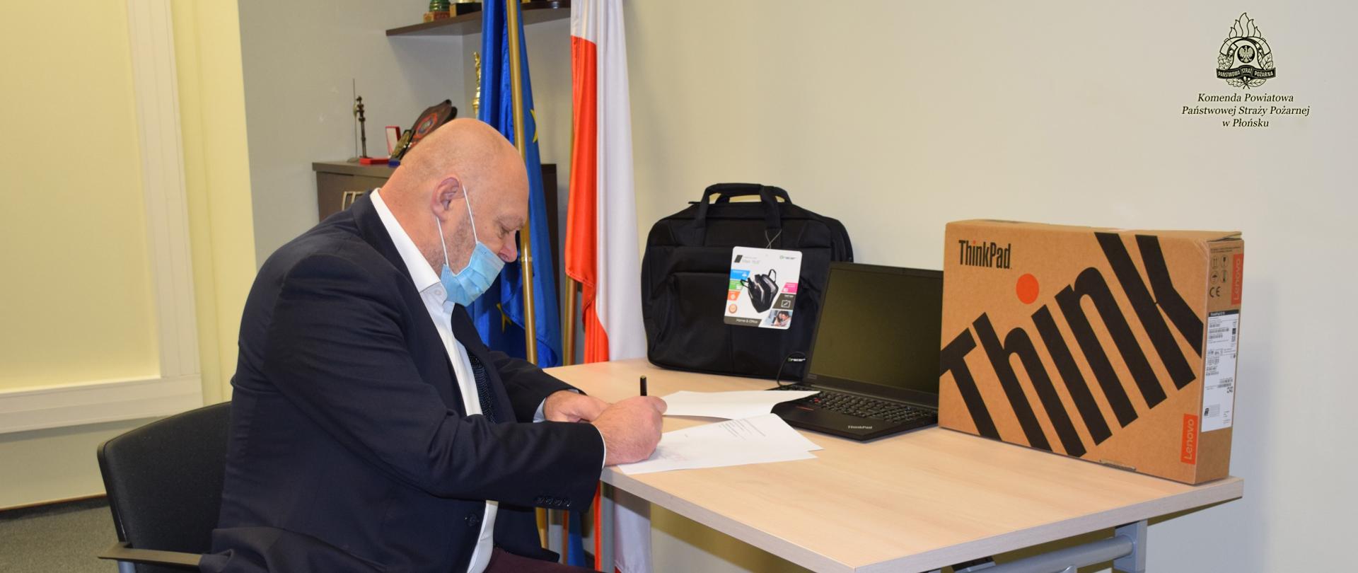 Mężczyzna podpisuje dokument, na stoliku stoi laptop, torba i pudełko, z tyłu półka z pucharami i flaga unijna. 