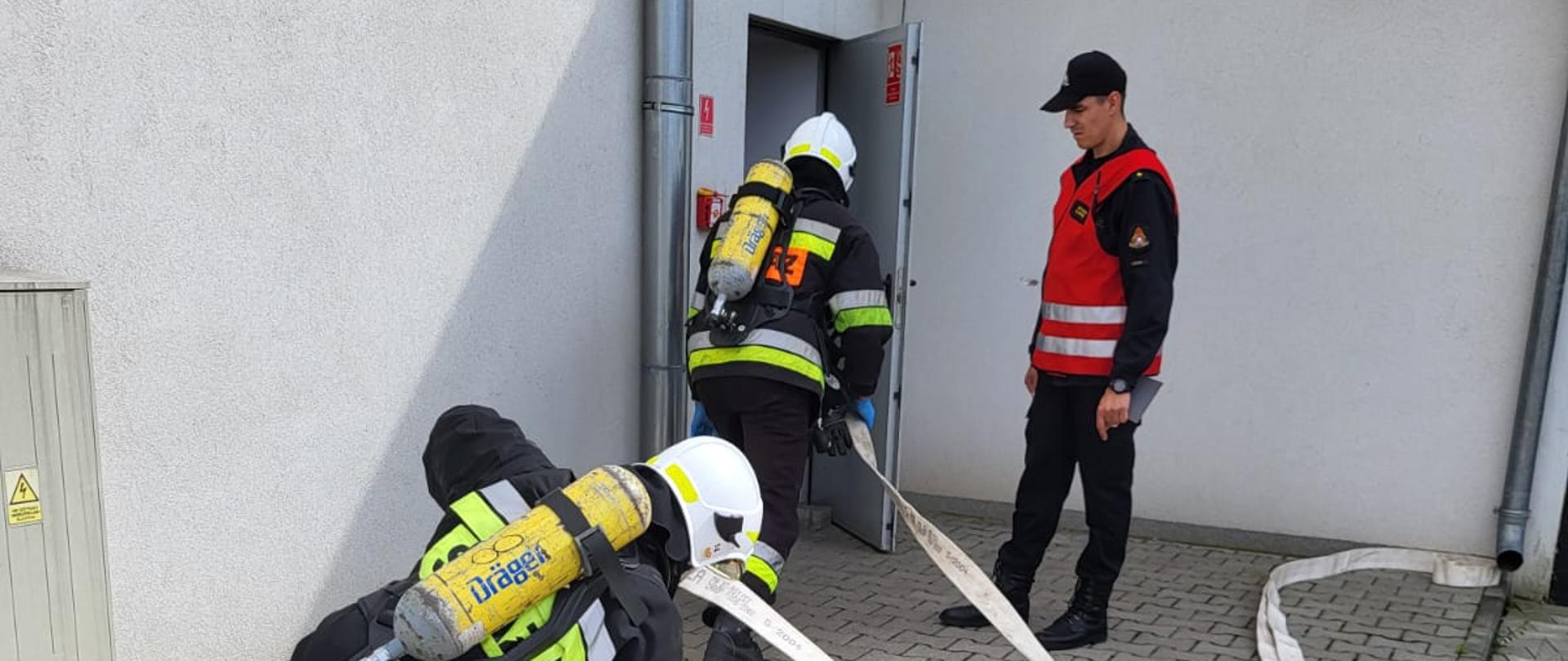 Strażacy w umundurowaniu bojowym rozwijają węże strażackie i wchodzą do pomieszczenia.