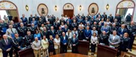Zdjęcie przedstawia dużą grupę około 80 osób, zebranych na uroczystości w przestronnej, bogato zdobionej sali sejmiku województwa śląskiego.Większość osób na zdjęciu to mężczyźni w ciemnych mundurach wyjściowych ochotniczej straży pożarnej z licznymi medalami i odznaczeniami. Wśród nich są również kobiety, niektóre w mundurach, inne w eleganckich strojach. Niektórzy mężczyźni są ubrani w garnitury. Grupa stoi na kilku poziomach. Na pierwszym planie, bliżej dołu zdjęcia, widać półokrągły, ciemny, drewniany stół, a przed nim stoją w rzędzie osoby, niektóre z nich trzymające niebieskie dyplomy. Osoby te są uśmiechnięte i patrzą w stronę obiektywu.Za nimi, na podwyższeniu, stoją kolejne rzędy ludzi, tworząc zwartą grupę. Większość z nich jest ustawiona tak, aby ich twarze były widoczne, a wiele osób uśmiecha się. Tło sali jest jasne i bardzo ozdobne. Ściany są wykonane z jasnego marmuru, z klasycznymi elementami architektonicznymi, takimi jak kolumny, pilastry, łukowate wnęki i zdobione gzymsy. We wnękach widać popiersia na postumentach. Górna część sali jest również bogato zdobiona, z rzeźbionymi detalami. Po prawej i lewej stronie widać wysokie okna, przez które wpada naturalne światło.
