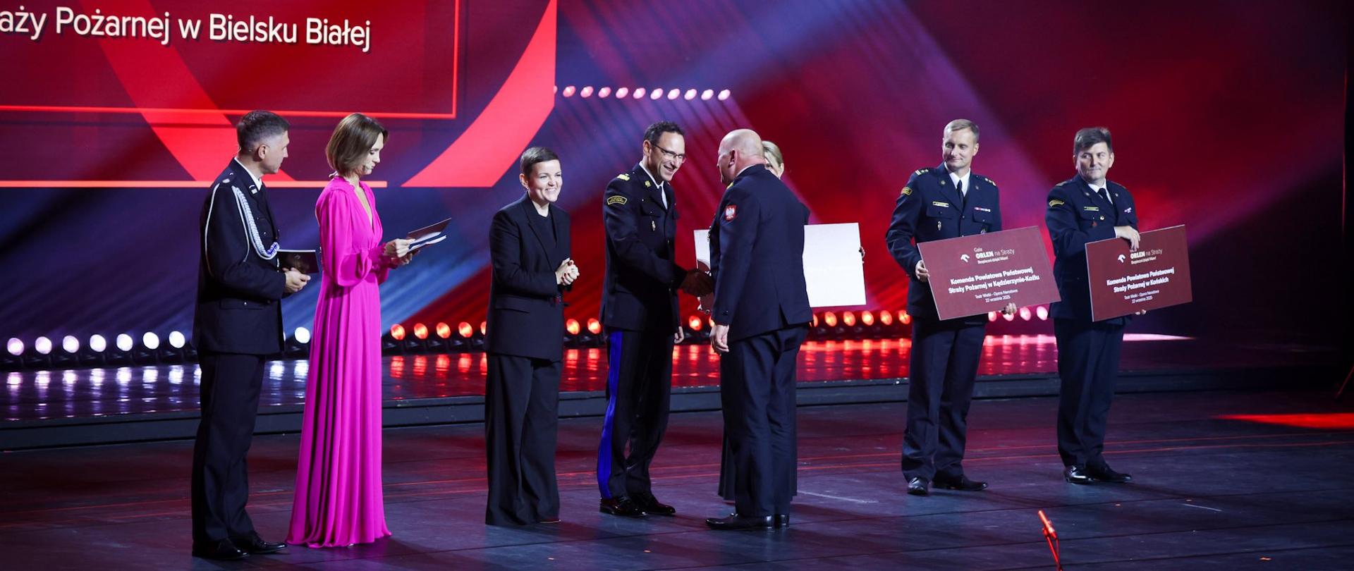  Zapytanie zostało przetworzone
Na scenie stoi sześć osób. Trzech mężczyzn w mundurach strażackich wymienia uścisk dłoni z mężczyzną w garniturze. Obok stoją dwie kobiety – jedna w czarnym, a druga w różowym stroju. W tle widoczny jest duży ekran, na którym wyświetlany jest tekst.