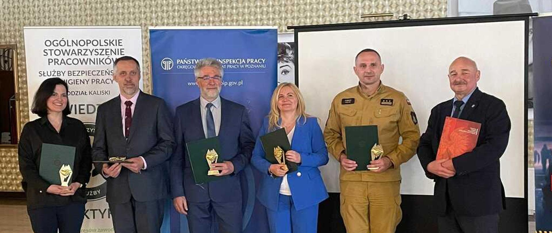 Zdjęcie w pomieszczeniu. Światło sztuczne. Widać osoby. Za osobami banery reklamowe i ekran. Na podłodze płytki. Na ścianie tapeta.