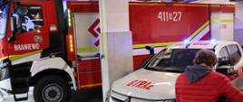 Zdjęcie zrobione w garażu bojowym PSP. Centralnie stoi duży czerwony samochód strażacki. Z prawej strony biały strażacki pikap.