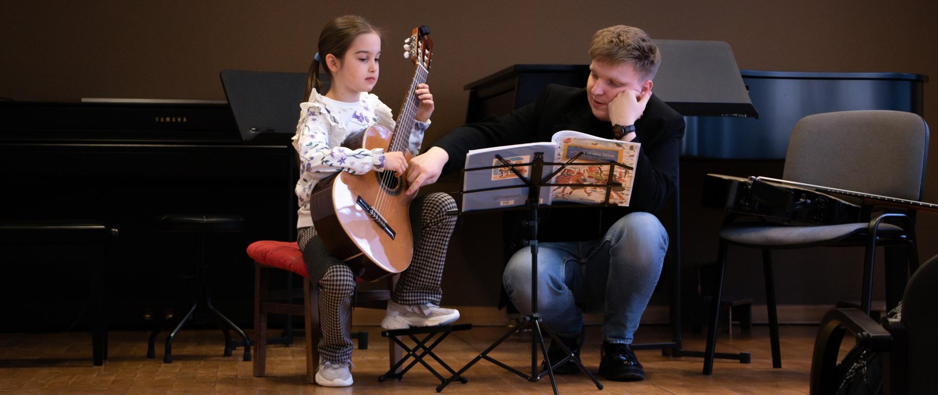 Prowadzący warsztaty w pozycji przykucnięcia wpatruje się w okolice otworu rezonansowego instrumentu, na którym gra młoda uczestniczka warsztatów. Wskazuje palcem na struny.