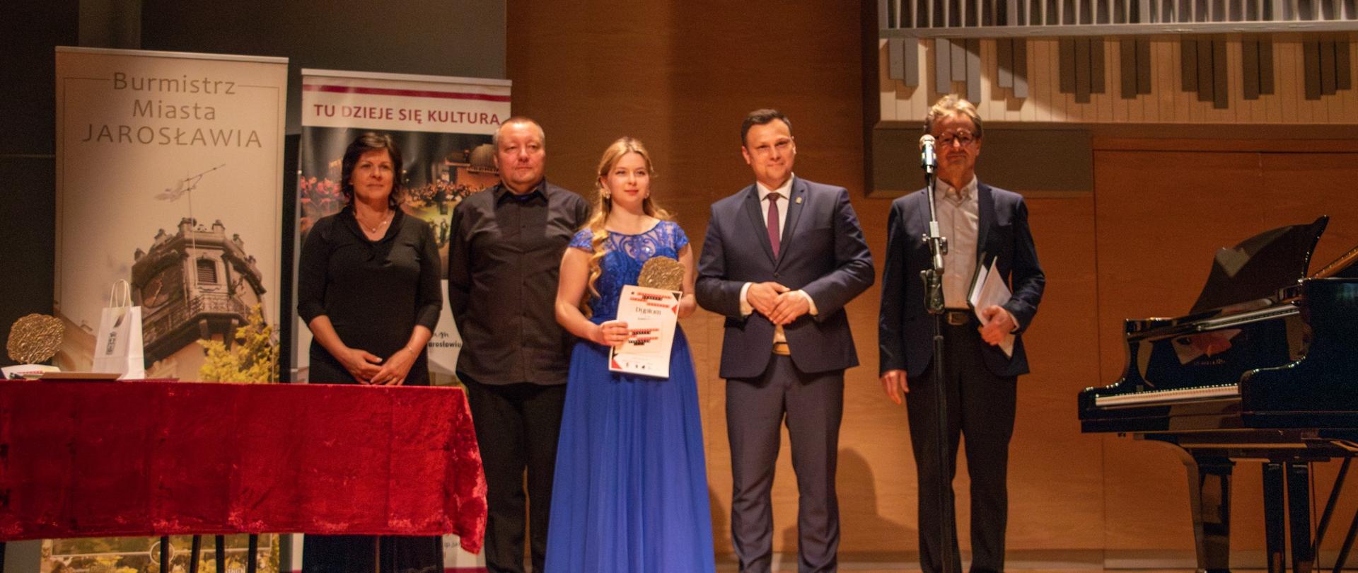 Zdjęcie przedstawia pięć osób stojących na scenie podczas ceremonii wręczenia nagród. Od lewej: kobieta w ciemnym stroju, mężczyzna w ciemnym ubraniu, kobieta w długiej, niebieskiej sukni z koronkowym gorsetem, trzymająca dyplom i nagrodę, mężczyzna w garniturze z krawatem oraz mężczyzna w okularach i garniturze, stojący przy mikrofonie i trzymający teczkę. Na scenie, po lewej stronie, widać stół z bordowym obrusem. Za stołem widoczny jest pionowy baner z napisem „Burmistrz Miasta Jarosławia”. Obok niego stoi kolejny baner z napisem „TU DZIEJE SIĘ KULTURA”. W tle, na ścianie, wiszą duże rury organowe, a nad fortepianem, po prawej stronie, wisi baner z tekstem w języku polskim, zawierającym słowa „OGÓLNOPOLSKI KONKURS MUZYKI POLSKIEJ” i „JAROSŁAW 2025”. Po prawej stronie zdjęcia widoczny jest również fragment czarnego fortepianu. Scena ma drewnianą podłogę.