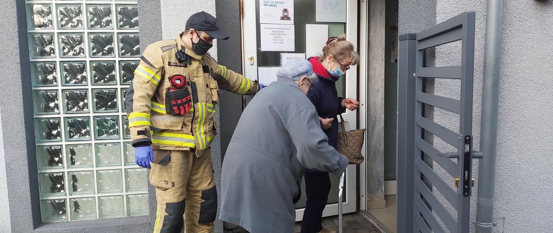 zdjęcie przedstawia strażaka który pomaga wraz z inną osobą starszej kobiecie wejść do punktu szczepień