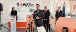 Mężczyzna w garniturze i w okularach stoi i mówi do kilku mikrofonów, za nim kilka osób i ścianka z napisem SOWA.