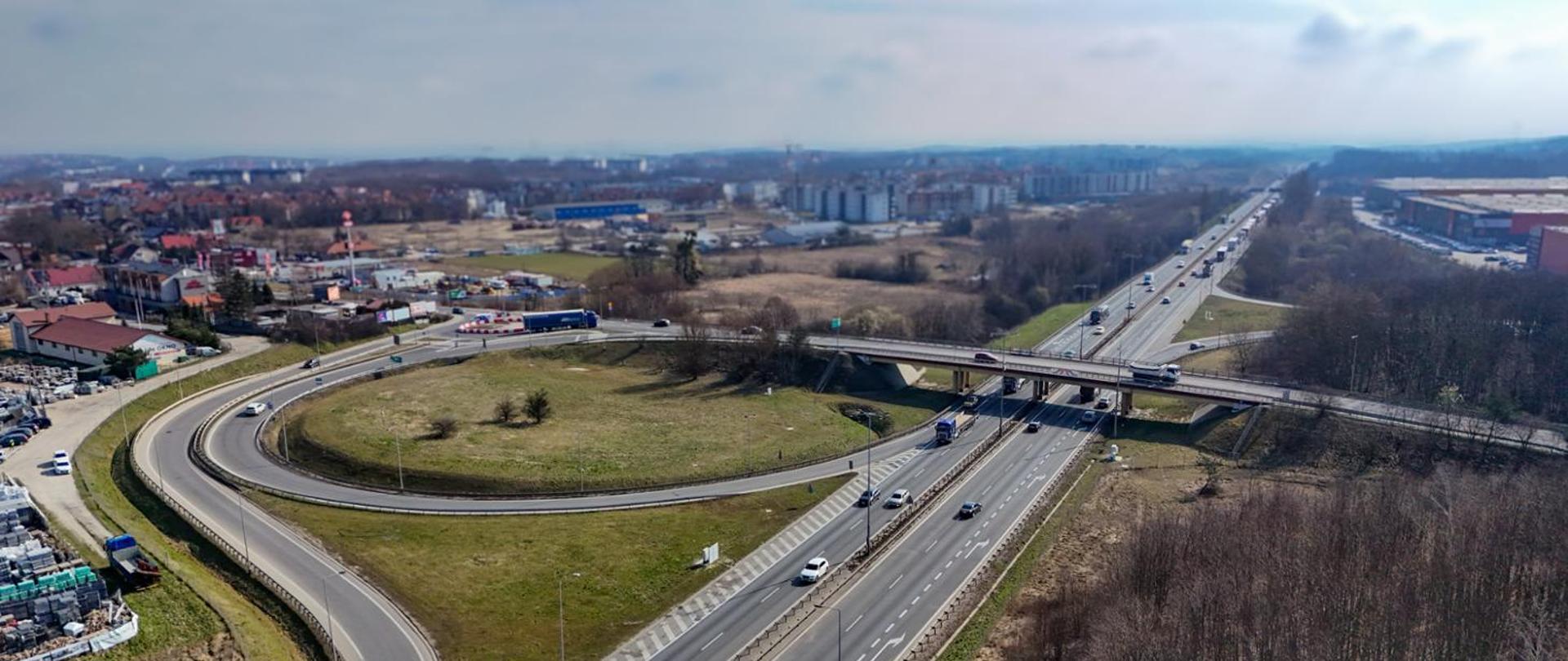 Widok z lotu ptaka na węzeł drogowy Kowale na Obwodnicy Trójmiasta. Na pierwszym planie widoczna jest duża pętla zjazdowa otaczająca trawiasty teren, nad wielopasmową drogą przebiega wiadukt. Po jezdniach poruszają się samochody ciężarowe i osobowe. W tle znajdują się zabudowania miejskie, tereny przemysłowe oraz zadrzewienia.