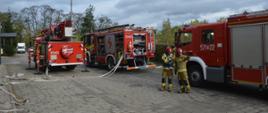 Ćwiczenia w ZOL. Zdjęcie przedstawia 2 strażaków na tle trzech wozów strażackich. Jeden z pojazdów to podnośnik hydrauliczny. Obydwaj mają na sobie ubrania specjalne koloru piaskowego i czerwone hełmy. Strażak stojący z lewej strony ma założoną kamizelkę biało-niebieską kierującego działaniem ratowniczym. W prawej ręce trzyma radiotelefon , a w lewej tabliczkę do monitorowania czasu pracy. Do pierwszego samochodu podłączone są dwa białe węże.Drugi strażak wskazuje na coś lewą ręką.