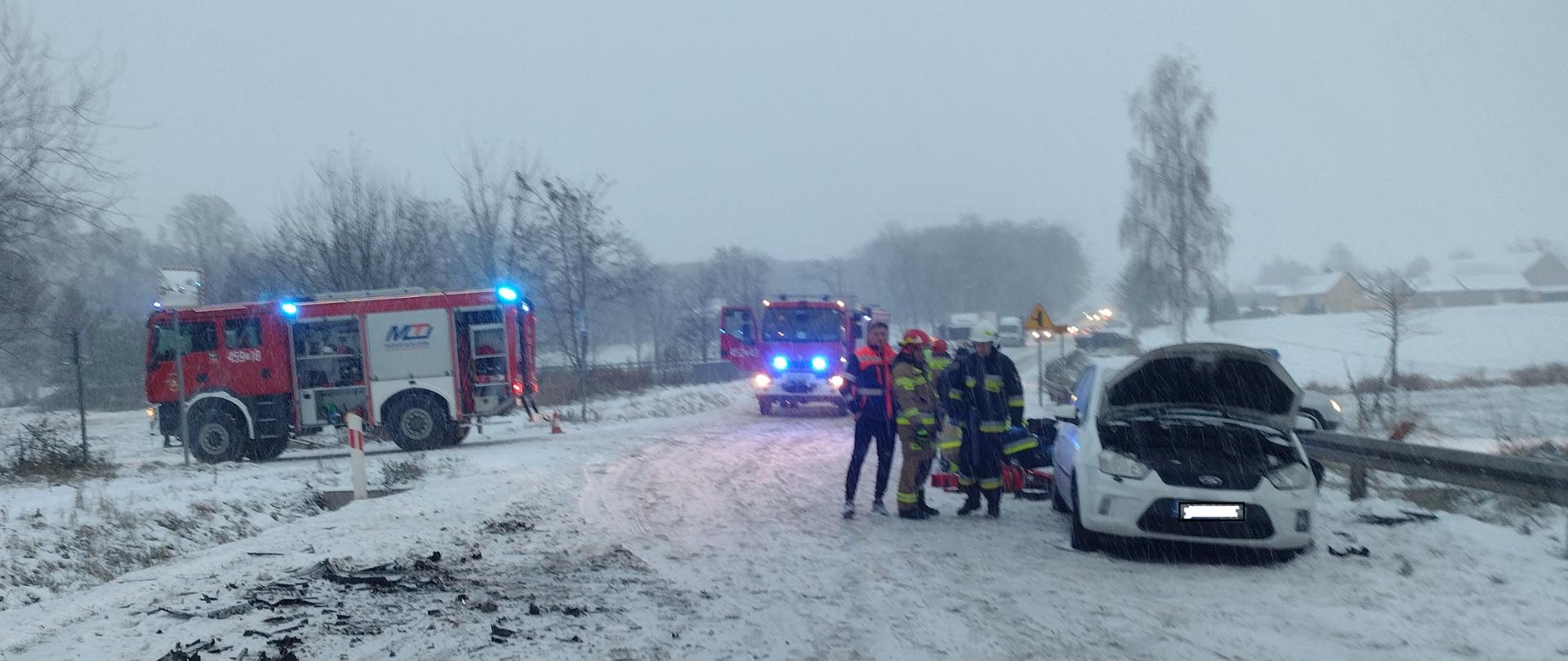 Na zdjęciu w godzinach popołudniowych w aurze zimowej na pierwszym planie widać ratoników i strażaków przy rozbitym aucie osobowym koloru białego. Jego maska jest podniesiona do góry. Na dalszym planie widać samochody gaśnicze na sygnałach oraz rozbite części aut osobowych lezące na jezdni. Zdjecie wykonane na drodze wojewódzkiej nr 754 w gm. Waśniów.
