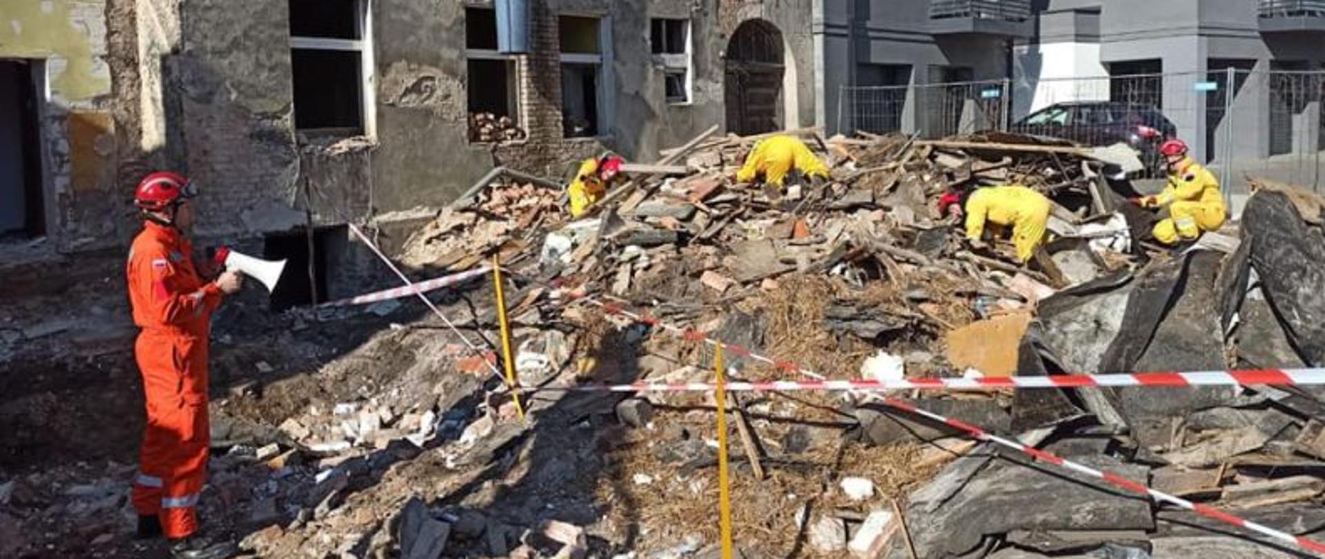 Grupa strażaków stoi na górze gruzów. Ubrani w czerwone hełmy i piaskowe mundury grupy poszukiwawczej. Od lewej strony stoi strażak z megafonem i kieruje ćwiczeniami.