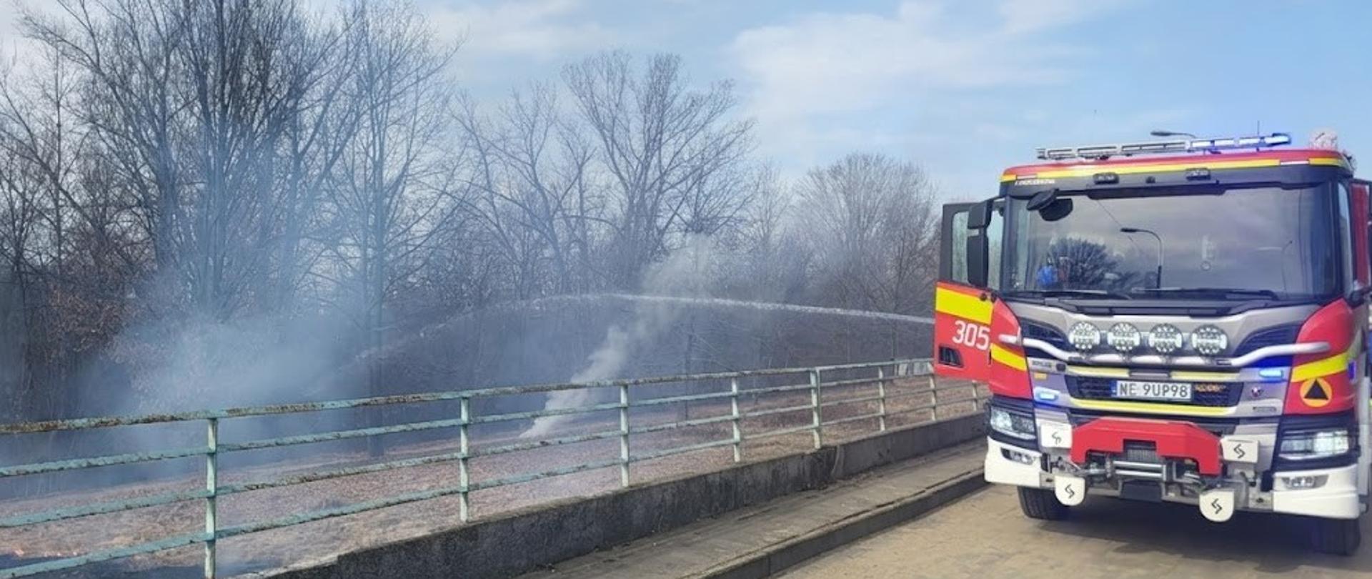 Pożar traw przy Wale Miedzeszyńskim. Strażacy apelują: „Stop pożarom traw!” Samochód straży pożarnej czerwony z żółtymi odblaskami. Widoczny strumień wody kierowany na dymiącą się trawę.