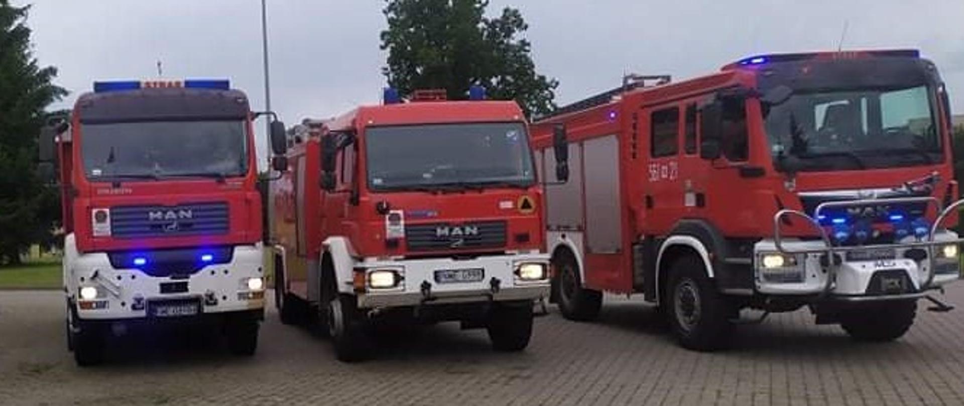 Na placu KP stoją strażacy i 3 samochody gaśnicze. Samochodu mają włączone światła błyskowe. Za strażakami widać budynek Komendy. 