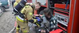 Pożar budynku mieszkalnego w Wydawach. Dwóch strażaków przy wozie strażackim udziela kwalifikowanej pierwszej pomocy mężczyźnie, który wskutek pożaru doznał poparzeń twarzy i ręki.