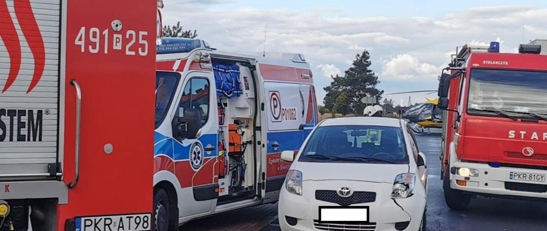 Zdjęcie przedstawia samochody służb pożarniczych oraz karetkę pogotowia ratunkowego. Na drodze stoi również samochód osobowy, który potrącił dwie osoby.