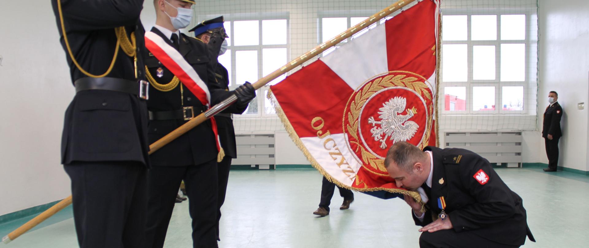Na zdjęciu widać młodszego brygadiera Wojciecha Pernala, który przyklęknął na czerwonym dywanie przed sztandarem i całuje sztandar. Sztandar Komendy Powiatowej Państwowej Straży Pożarnej w Łęczycy jest pochylony. Na zdjęciu widać strażaków w mundurach wyjściowych. Na twarzach strażacy mają założone białe maseczki. Przez okna widać, że jest dzień.