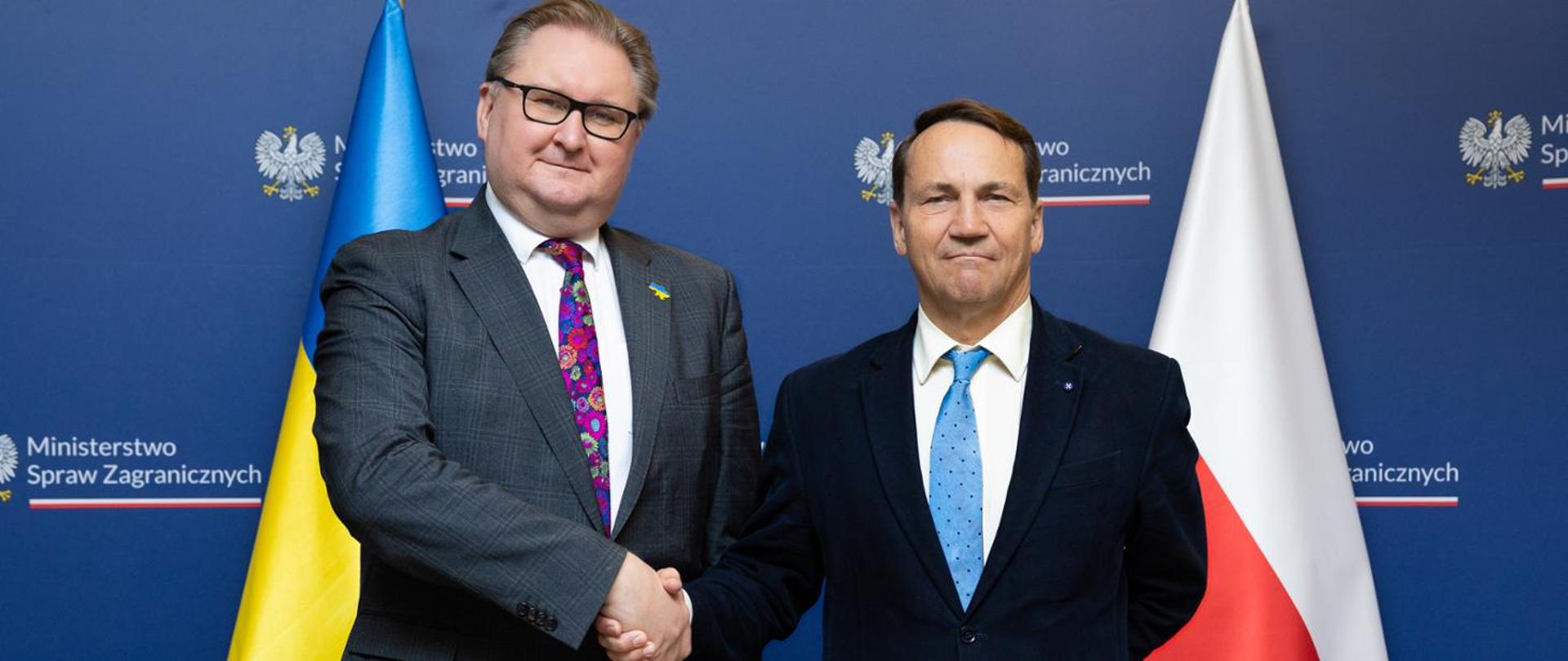 Na zdjęciu jest dwóch mężczyzn. Podają sobie dłonie. Mają na sobie garnitury. Stoją na tle niebieskiej ścianki z napisem Ministerstwo Spraw Zagranicznych i dwóch flag - Polski i Ukrainy. 
