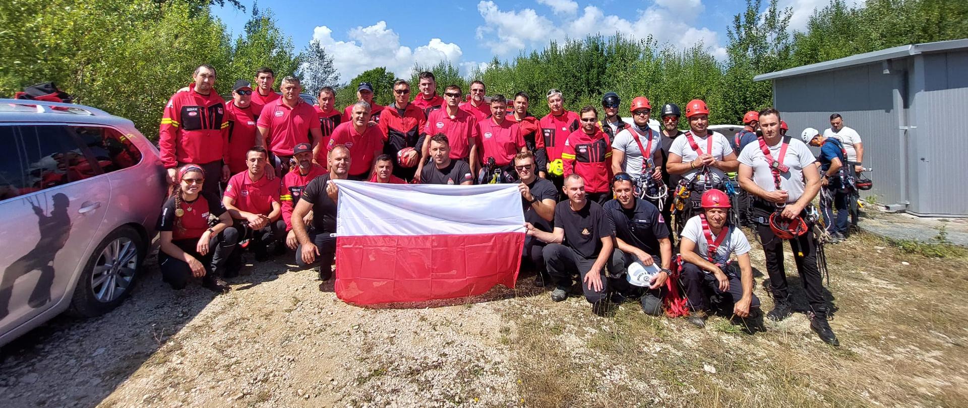 Na zdjęciu widoczna grupa strażaków w czerwonych i szarych koszulkach z Polski oraz Rumunii. Jest to wspólne zdjęcie podczas warsztatów ratowniczych z zakresu ratownictwa wysokościowego. Ratownicy po środku grupy trzymają biało-czerwoną flagę.