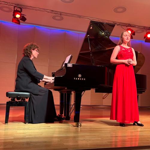 Zdjęcie przedstawia uczennicę podczas występu wokalnego z towarzyszeniem akompaniatorki grającej na fortepianie na scenie sali koncertowej, w tle baner szkoły