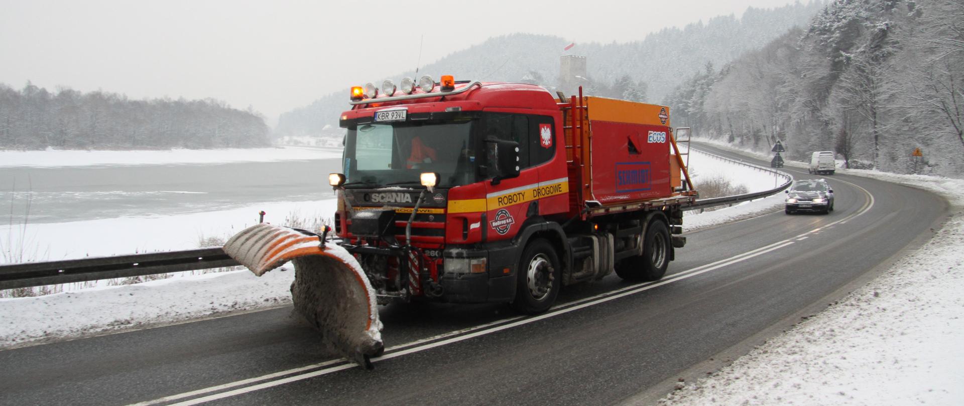 Zimowe utrzymanie DK75, okolice zamku Tropsztyn w woj. małopolskim. Na pierwszym planie pługosolarka w akcji na drodze, w tle zimowy krajobraz. 