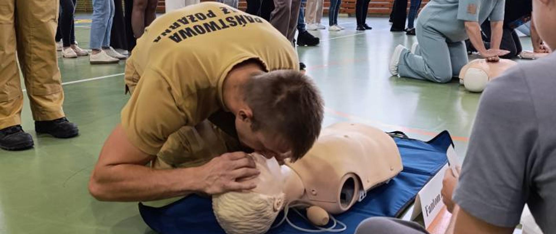 Zdjęcie przedstawia strażaka wykonującego resuscytację krążeniowo oddechową na fantomie, za nim stoją osoby oczekujące na swoją kolej w prowadzeniu resuscytacji