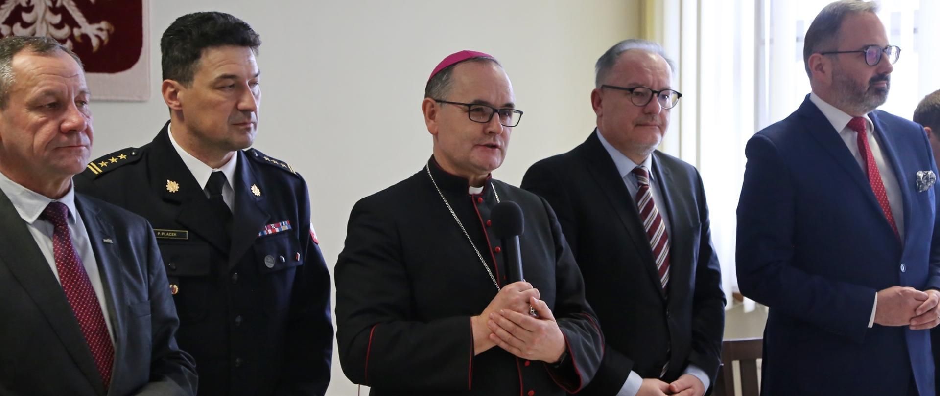 Ksiądz stojąc z mikrofonem za stołem błogosławi, obok stojący pozostali goście oraz Komendant CS PSP