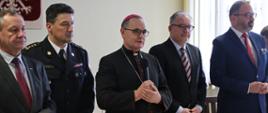Ksiądz stojąc z mikrofonem za stołem błogosławi, obok stojący pozostali goście oraz Komendant CS PSP