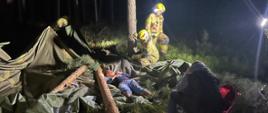 Strażacy z Ochotniczej Straży Pożarnej w ubraniach bojowych docierają na miejsce zdarzenia, gdzie przymierzają się do udzielenia pierwszej pomocy poszkodowanemu na terenie zerwanego obozowiska w lesie.