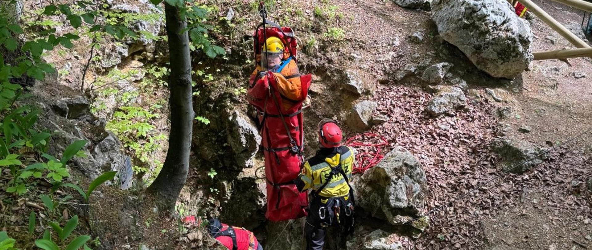 na zdjęciu poszkodowany przypięty do noszy wyciągany na linie pionowo w górę z jaskini obok niego dwóch ratowników
