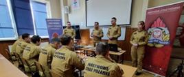Sala szkoleniowa – przodem stoi komendant powiatowy PSP i strażacy z KW PSP Kraków prowadzący zajęcia, tyłem widoczni siedzący uczestnicy szkolenia