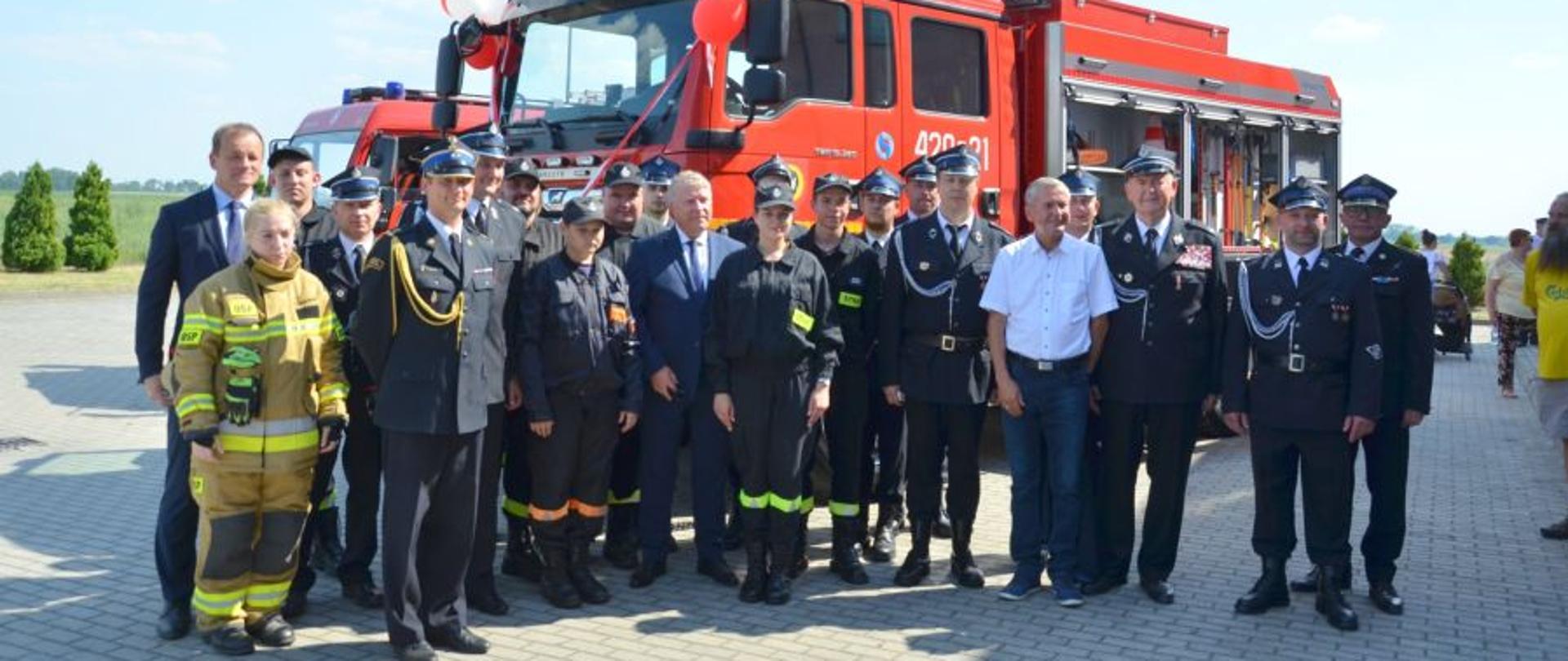 Grupa strażaków i osób cywilnych pozuje na tle nowego czerwonego samochodu strażackiego