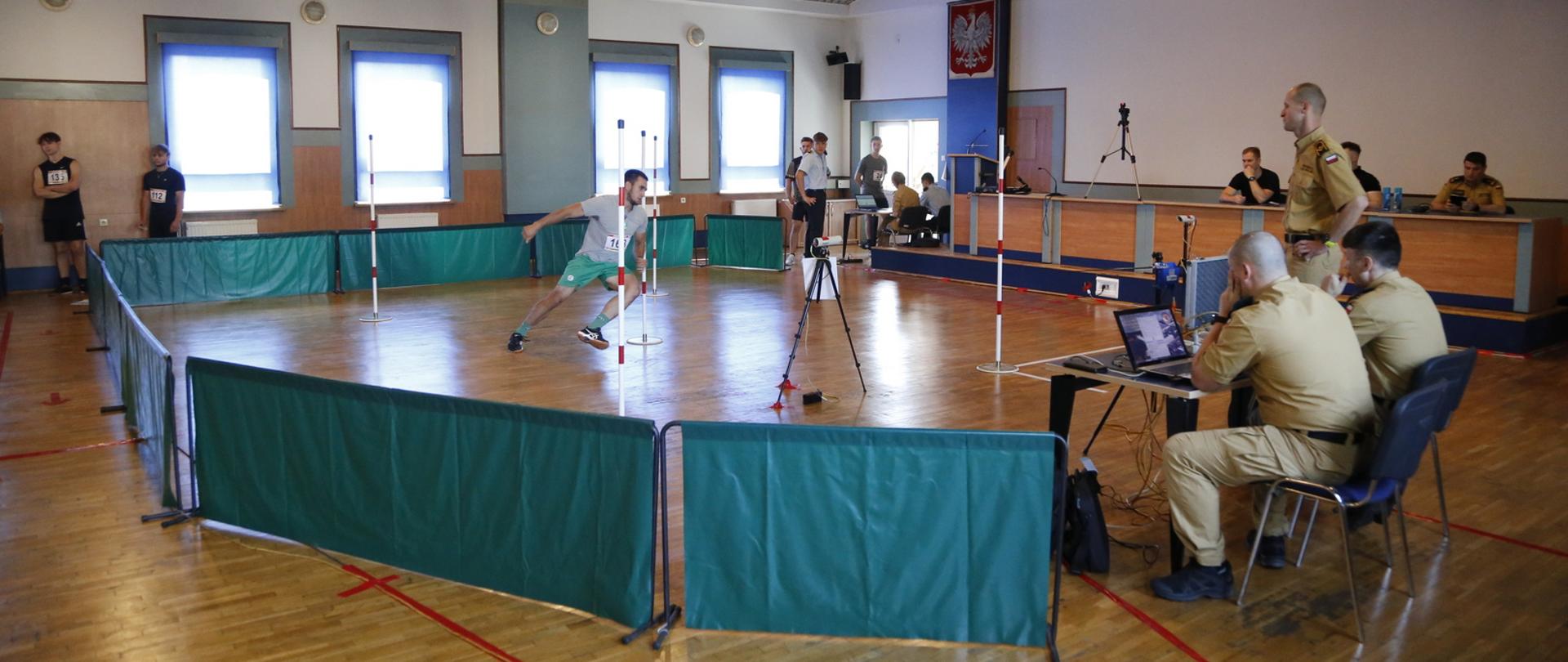 Po prawej dwaj strażacy siedzący przy ławie z urządzeniami do pomiaru czasu przed nimi kandydat uczestniczący w konkurencji bieg po kopercie