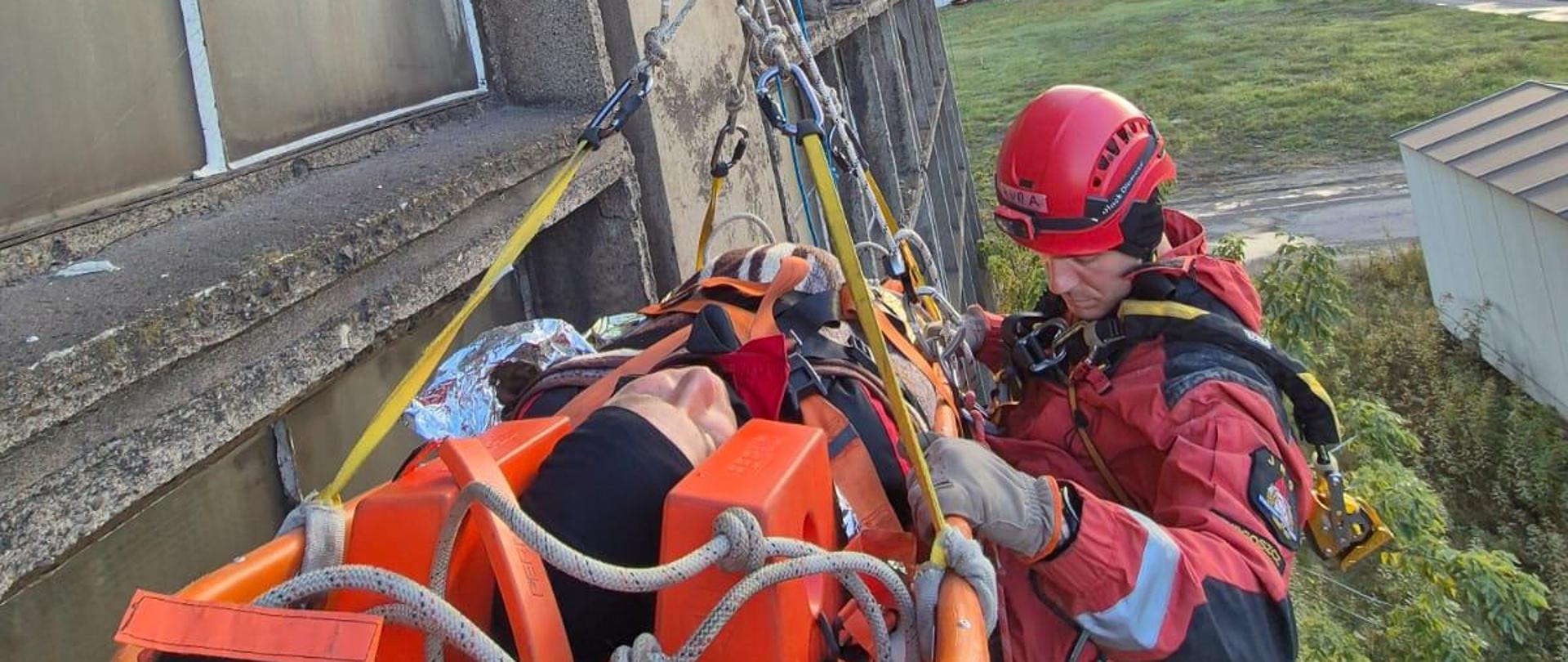 Ratownik w czerwonym kombinezonie i kasku opuszcza na linach osobę w noszach ratowniczych wzdłuż ściany betonowego budynku. W tle widoczne tereny przemysłowe i zabudowania.