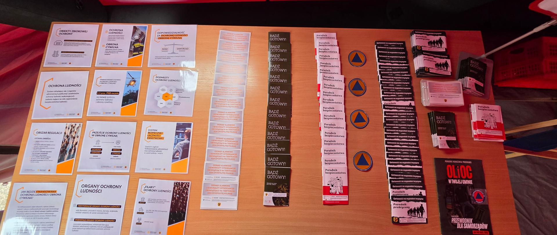 Na stole wyłożone są broszury, ulotki i plakaty dotyczące ochrony ludności, obrony cywilnej oraz przygotowania na sytuacje kryzysowe.