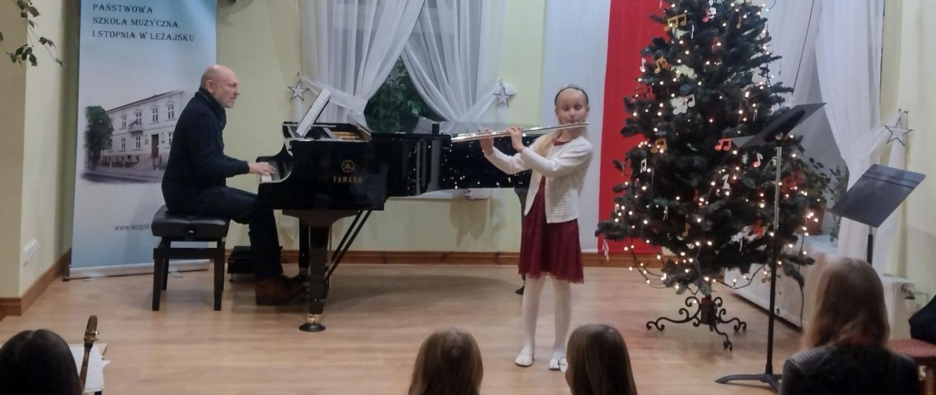 Zdjęcie z koncertu fletowego. Uczennica odświętnie ubrana gra na flecie. Za nią z tył po lewej strony (perspektywa oglądającego) czarny fortepian, przy nim siedzi akompaniator. Po prawej stronie udeorowana choinka. Zdjęcie w kolorach jasnych.