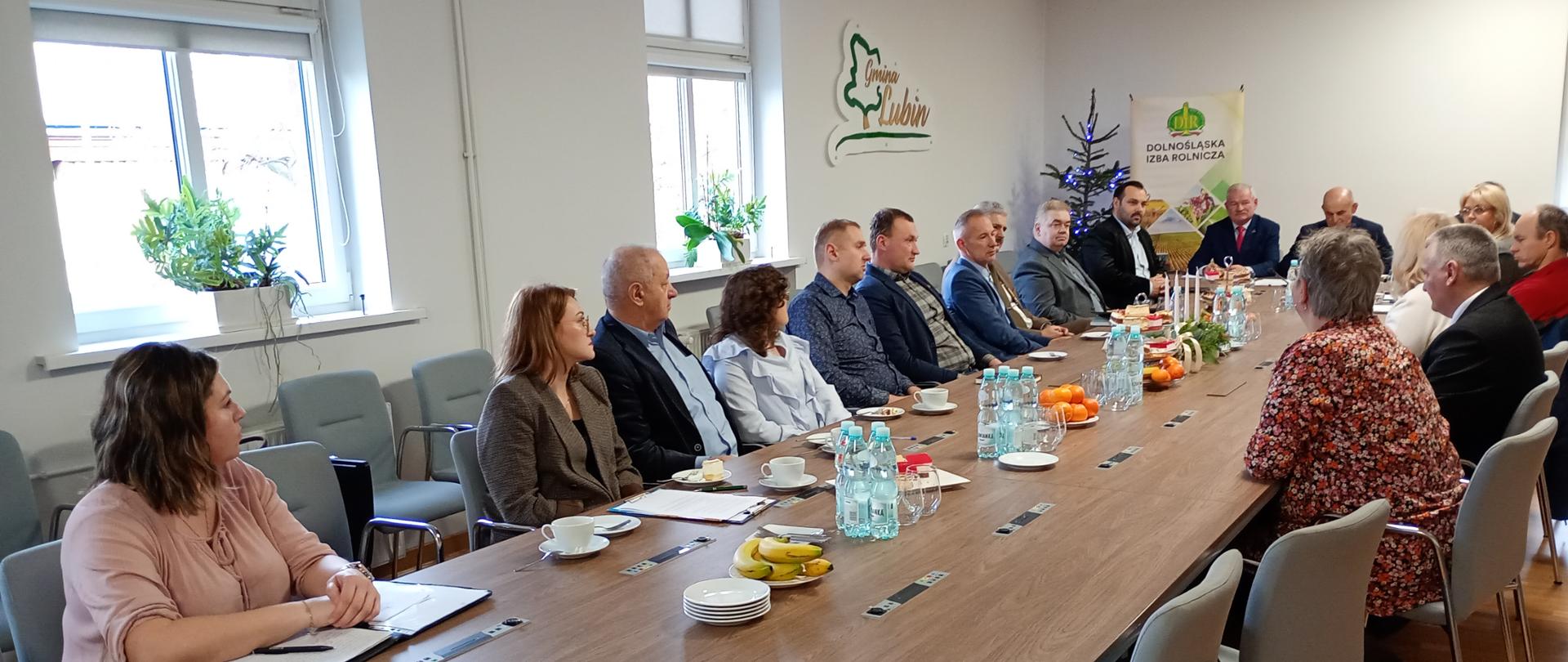 Na zdjęciu przedstawiono spotkanie członków Rady Powiatowej Dolnośląskiej Izby Rolniczej powiatu lubińskiego. Uczestnicy siedzą przy długim stole w sali konferencyjnej. W tle widoczna jest dekoracja z logo "Gmina Lubin". Na stole znajdują się napoje, owoce, a także notesy i długopisy.