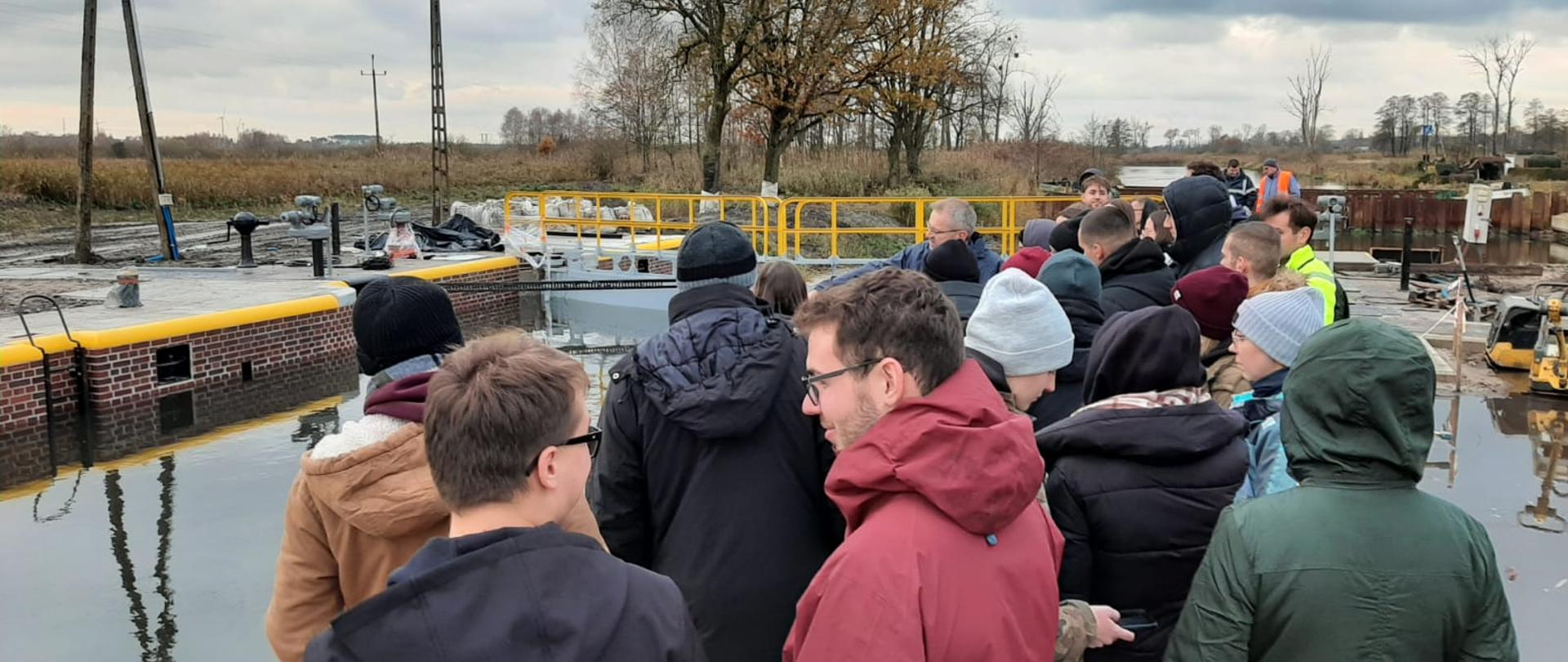 Studenci i pracownicy Uniwersytetu Przyrodniczego w Poznaniu na placu budowy remontowanej śluzy Nakło Wschód. Postaci pierwszoplanowe odwrócone tyłem do obiektywu aparatu fotograficznego zwróceni ku komorze śluzy napełnionej wodą i słuchający Kierownika Nadzoru Wodnego w Nakle nad Notecią opowiadającego o fazach wykonywanego remontu. W tle rozstawione maszyny budowalne oraz pracujący w kaskach budowniczowie. 