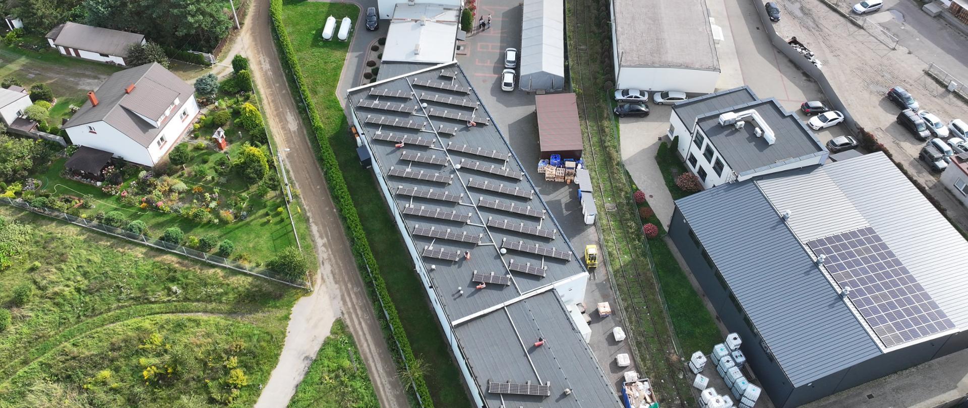 Zdjęcie zakładu przemysłowego z powietrza. Kompleks kwadratowych budynków z instalacją wyciągowa i panelami słonecznymi na dachach. Pomiędzy budynkami stoją samochody. Na prawo do zakładu jest parking, na lewo dwa domy jednorodzinne otoczone ogrodami.