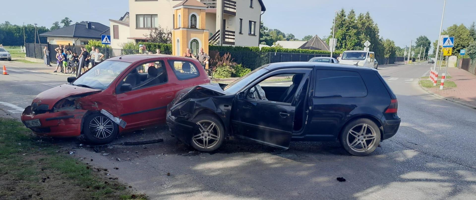 Na zdjęciu widoczne są dwa pojazdy osobowe biorące udział w zdarzeniu, znajdują się one na skrzyżowaniu. Oba pojazdy mają zniszczony przód w wyniku uderzenia.