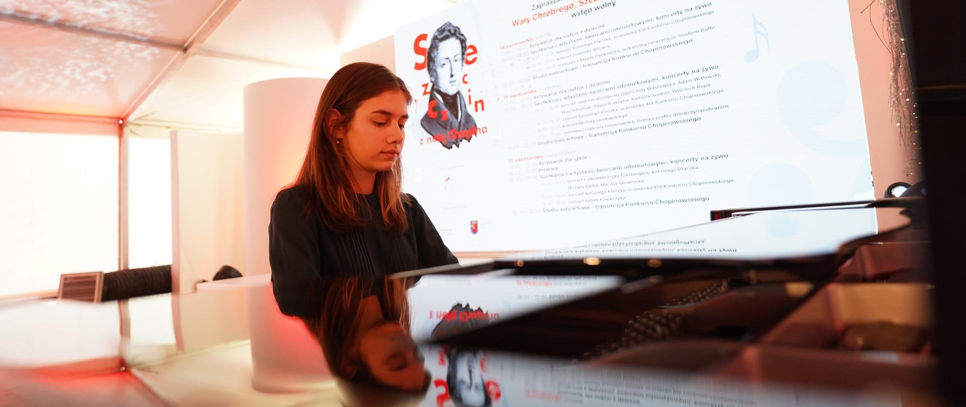 zdjęcie przedstawia uczennicę grającą na fortepianie na tle ekranu multimedialnego z logo sponsora koncertu